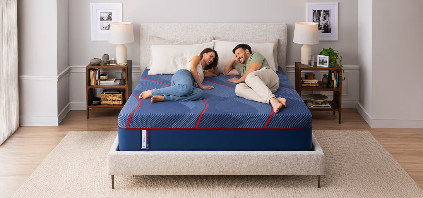 Man and woman lying on a blue mattress in a bedroom with white walls and wooden floors.