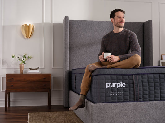 Man sitting on a luxury Purple ultimate mattress holding a mug in a room with a side table and decorative items.
