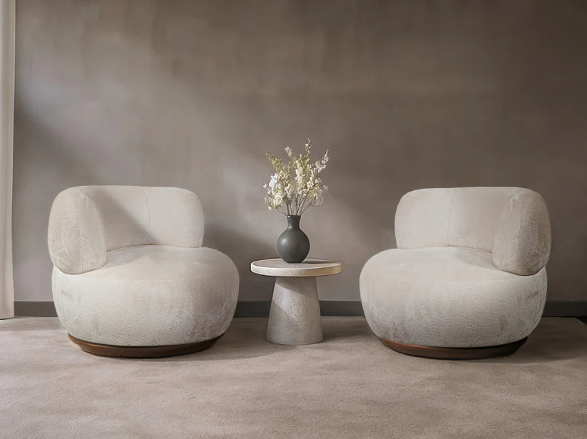 Two beige armchairs with a small round table and vase of flowers against a plain wall.