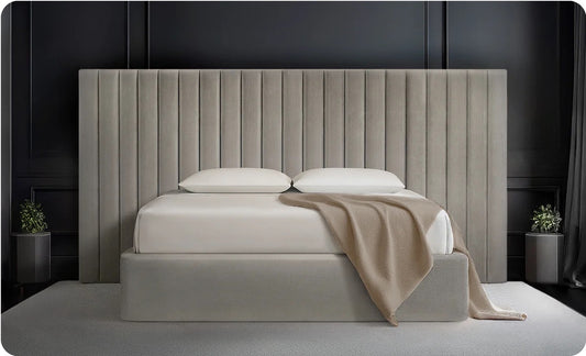 Beige upholstered bed with white bedding and a beige blanket against a dark wall.