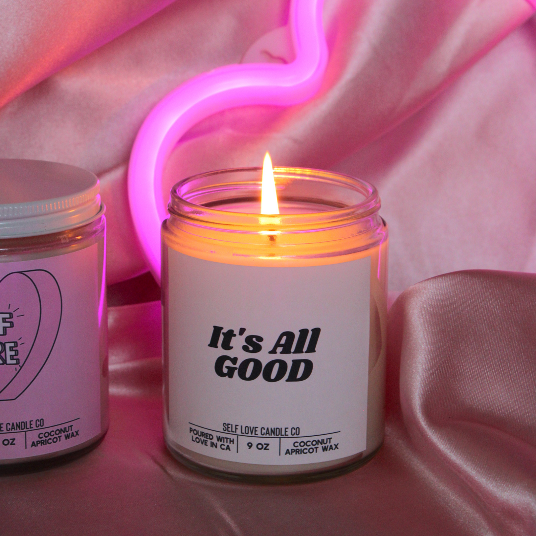 Candle in a jar labeled 'It's All Good' with a neon light in the background