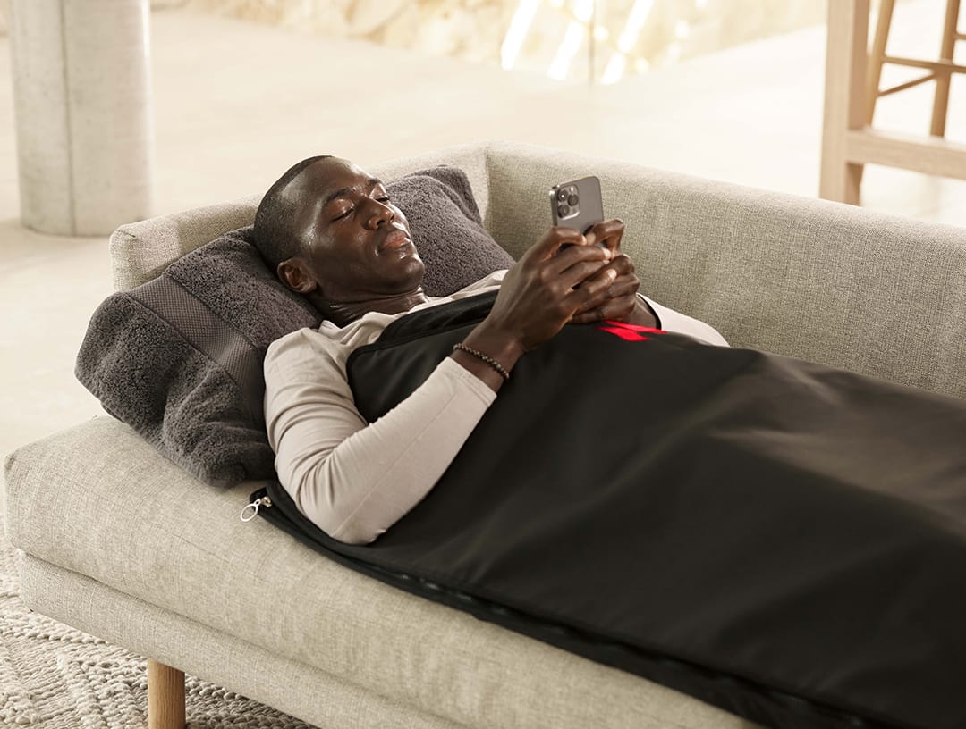 Man lying on a couch using a smartphone in a black sauna blanket.