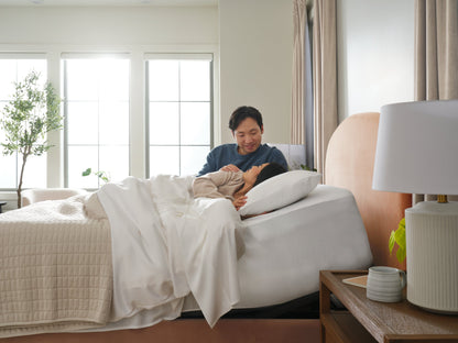 Two people lying in bed together in a bright bedroom with large windows.