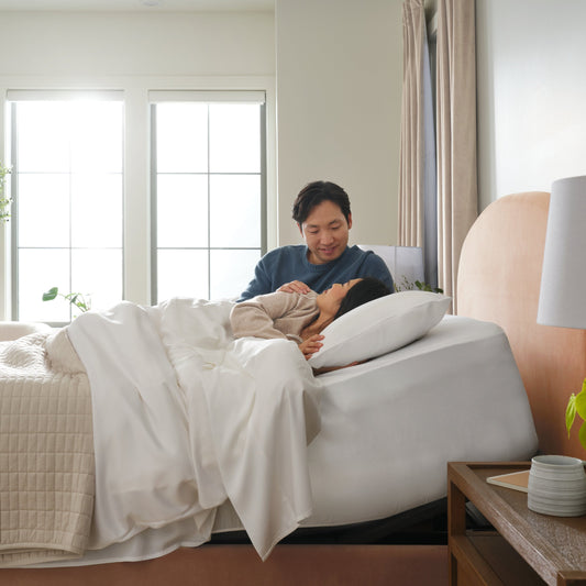 Two people lying in bed together in a bright bedroom with large windows.