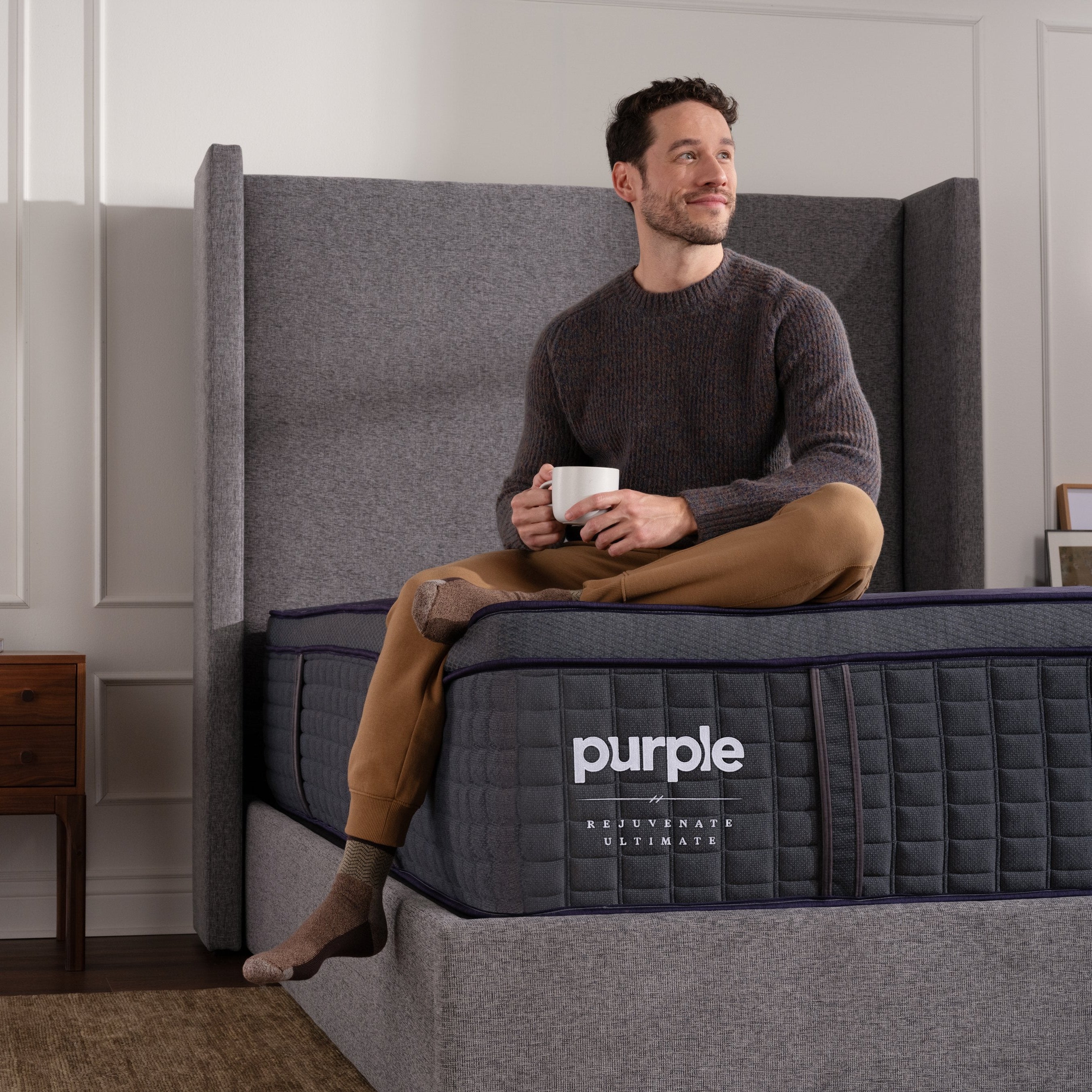 Man sitting on a luxury Purple ultimate mattress holding a mug in a room with a side table and decorative items.