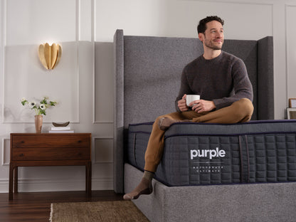 Man sitting on a luxury Purple ultimate mattress holding a mug in a room with a side table and decorative items.