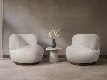 Two beige armchairs with a small round table and vase of flowers against a plain wall.