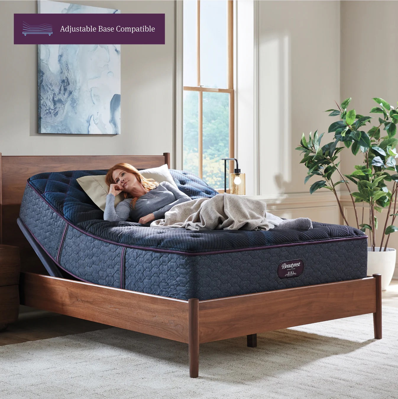 Woman lying on a blue mattress in a bedroom with adjustable base compatibility text.