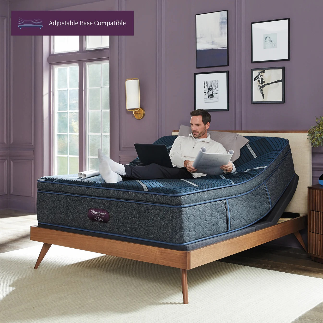 Man using a laptop on a adjustable bed with 'Beautyrest' branding in a room with purple walls and large windows.