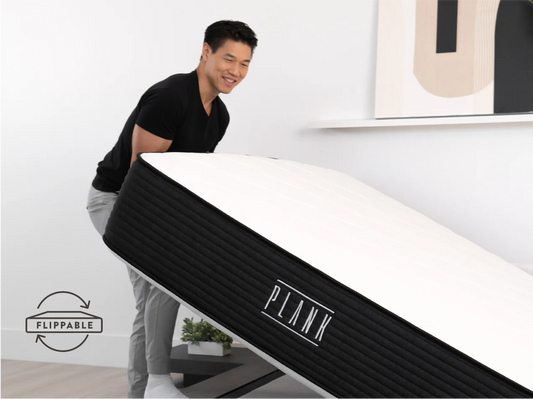 Person interacting with a black and white bed frame labeled 'Plank' in a minimalistic room.