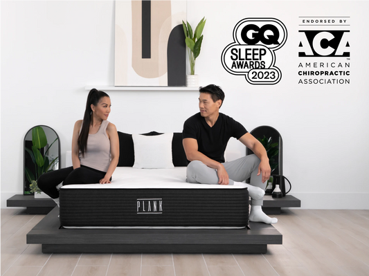 Two people sitting on a PLANK mattress with GQ Sleep Awards 2023 and ACA logos in the background.