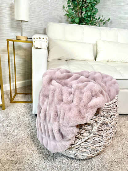 Pink fluffy blanket in a wicker basket on a white couch with a lamp and plant in the background.