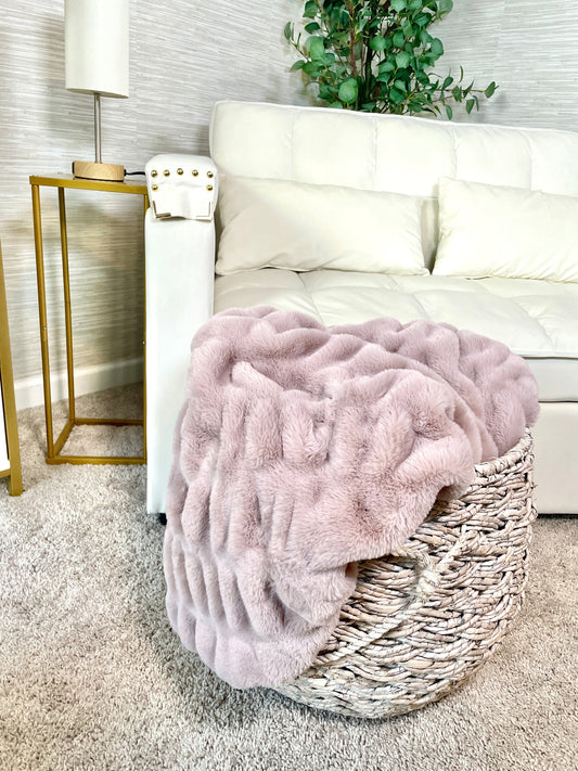 Pink fluffy blanket in a wicker basket on a white couch with a lamp and plant in the background.
