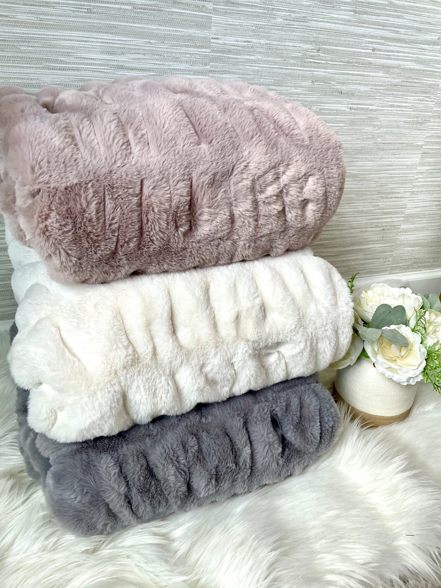Stack of three textured blankets in pink, white, and gray on a fluffy surface with flowers.
