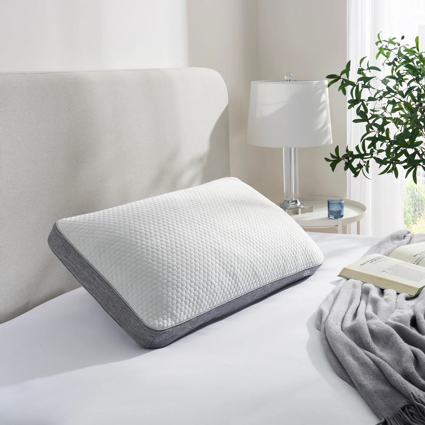 Ergonomic pillow on a bed in a bedroom setting with a lamp and plant.