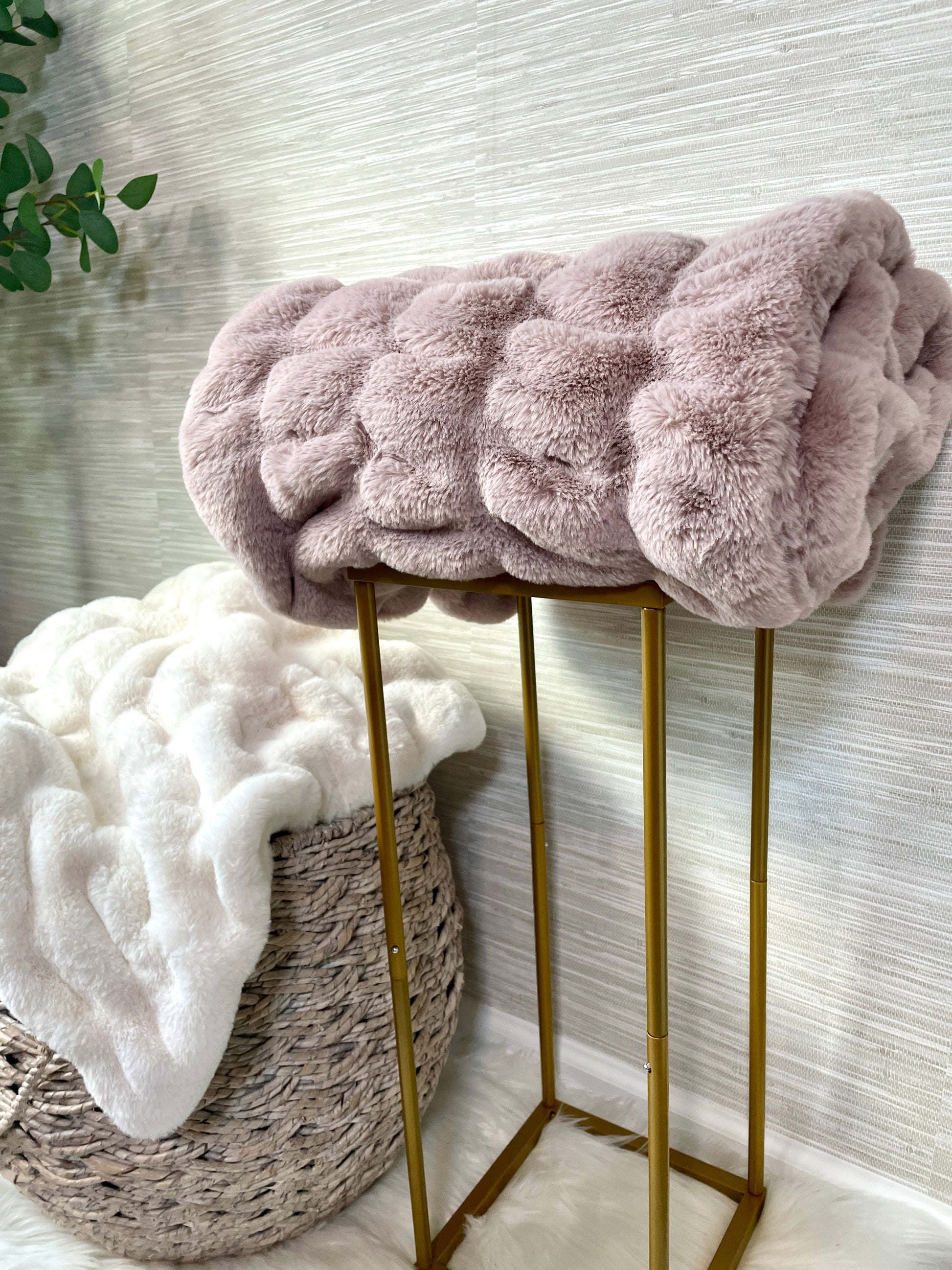 Pink fluffy blanket draped over a gold stool with a white textured wall in the background.