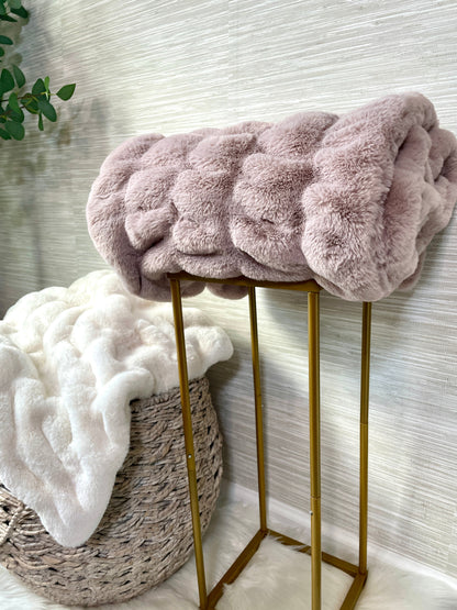 Pink fluffy blanket draped over a gold stool with a white textured wall in the background.