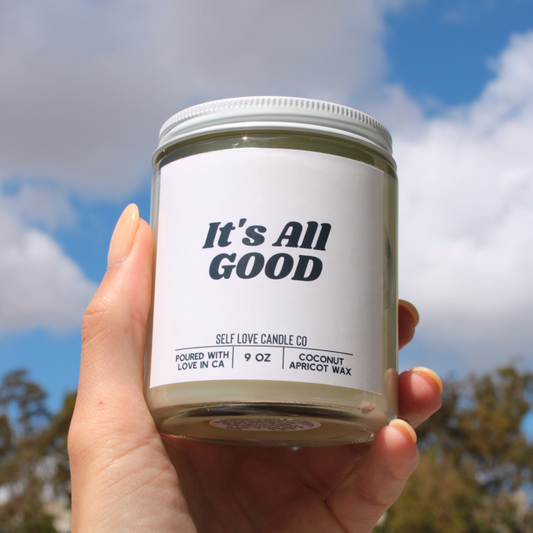 Hand holding a candle labeled 'It's All Good' against a blue sky background