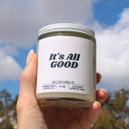 Hand holding a candle labeled 'It's All Good' against a blue sky background