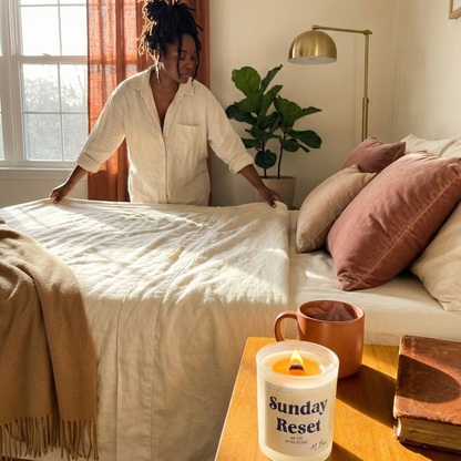 Woman making a bed in a cozy bedroom with a 'Sunday Reset' candle on a nightstand.