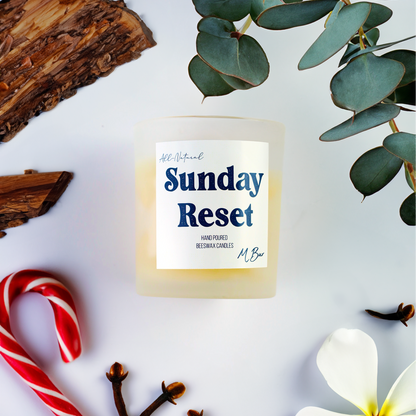 Candle labeled 'Sunday Reset' surrounded by eucalyptus, candy canes, and flowers on a white background