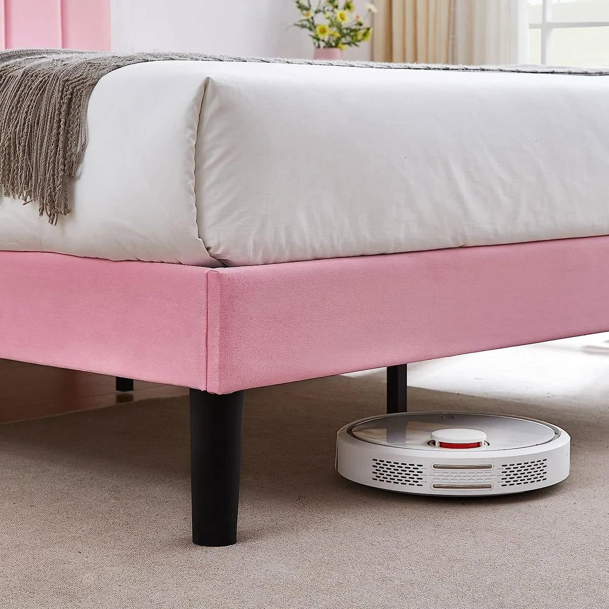 Robotic vacuum cleaner on a carpeted floor in front of a pink bed.