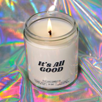 Candle in a glass jar labeled 'It's All Good' on a reflective, multicolored background